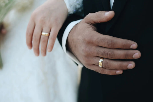 A close-up of a newlywed couple’s hands, each wearing a golden tungsten wedding band. The groom in a black suit and the bride in a white gown gently touch hands, symbolizing love and unity.